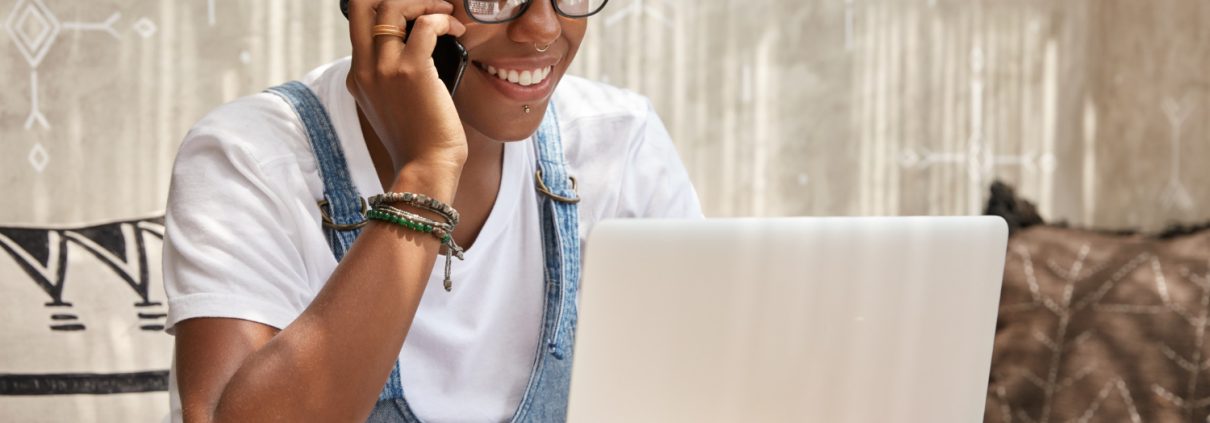 stylish-african-american-woman-calls-smart-phone-looks-laptop-computer-updates-software